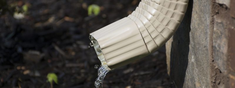 A rain gutter downspout with water coming out from the side, with a flower bed in the background. Shallow Depth of Field.