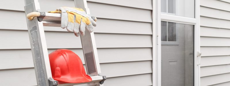 Red hard hat and work gloves on ladder with house siding background.